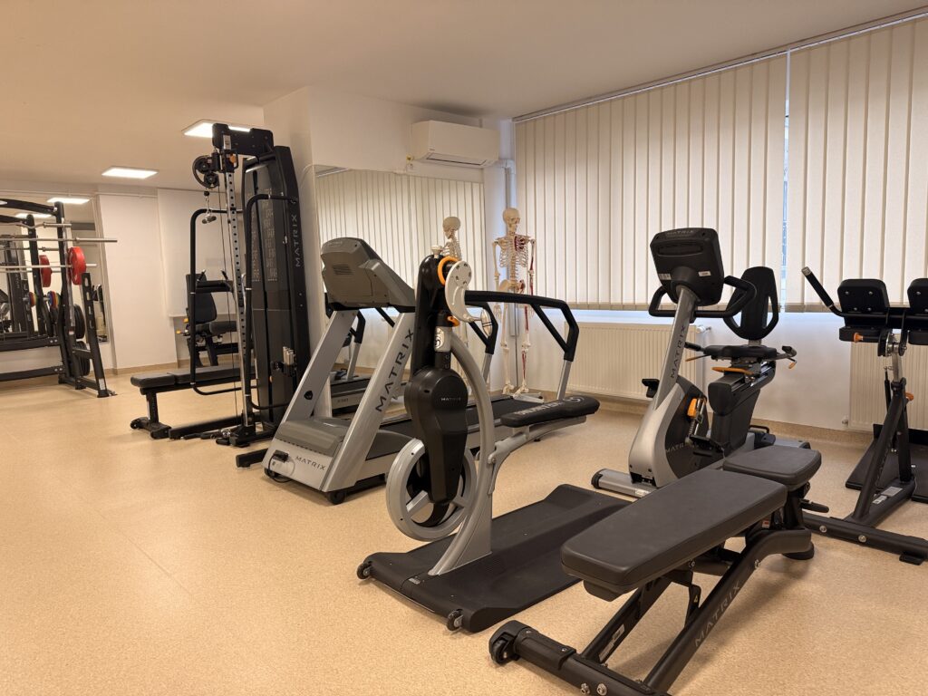 Poîă cu aparatele din sala de kinetoterapie alineate, bicicletă de mâini, bicicletă ortopedică, bandă de alergat cu înclinare dublă, scripete și suportul de siguranță pentru genoflexiuni cu bara.
